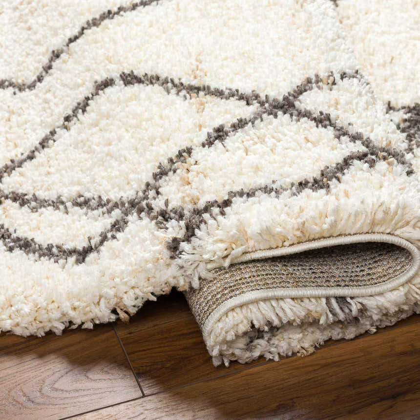 Close-up of the Hersz Cream Area Rug showing its shag texture and geometric brown line pattern on a wood floor. Fold