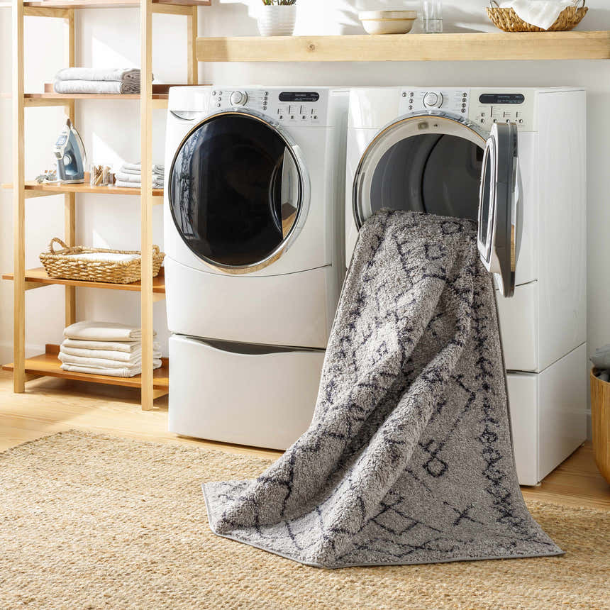 Laundry room scene with Elyes Gray Washable Area Rug coming out of the dryer, jute rug, wood shelves, and white appliances. Washing-Machine