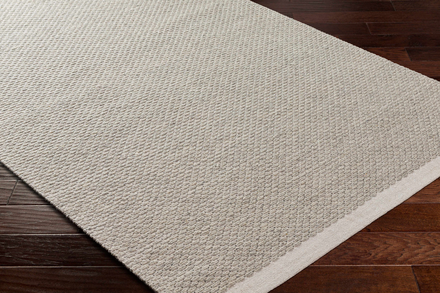 Close-up of the Light Brown Emlen Area Rug showing its woven texture and cream border on dark wood flooring. Corner