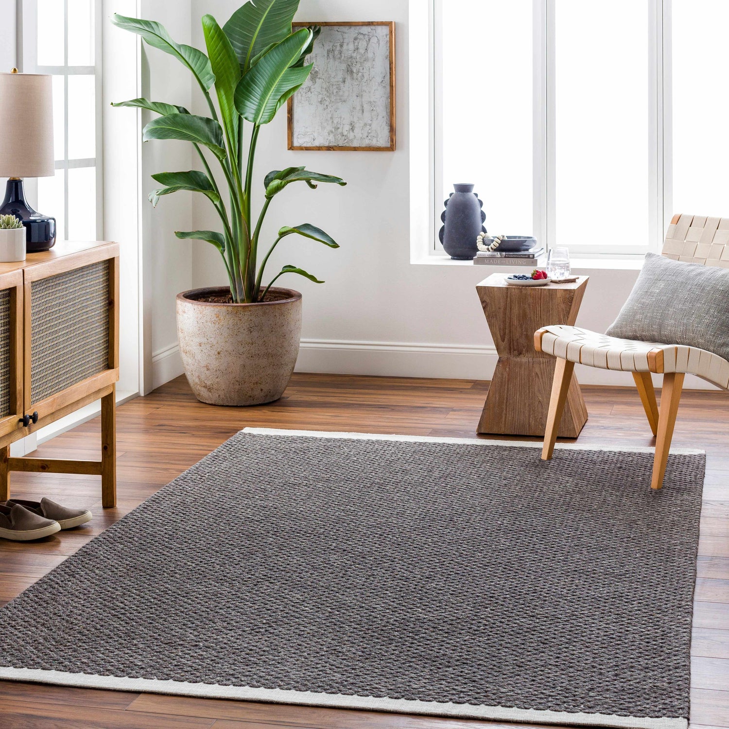 Living room scene with Emlen Nutmeg Area Rug, wood floors, woven chair, and potted plant near a bright window. Roomscene-Rectangle