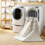 Laundry room scene with a gray and white geometric Cili Washable Area Rug partially inside a washing machine. Jute rug underneath. Washing-Machine