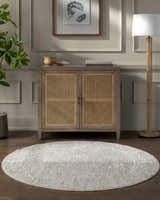 Living room scene with a light gray, round Kyran Area Rug under a cabinet with cane doors. Roomscene-Round