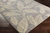 Close-up of the Snook Area Rug showing beige leaf pattern on a gray background, displayed on dark wood flooring. Corner