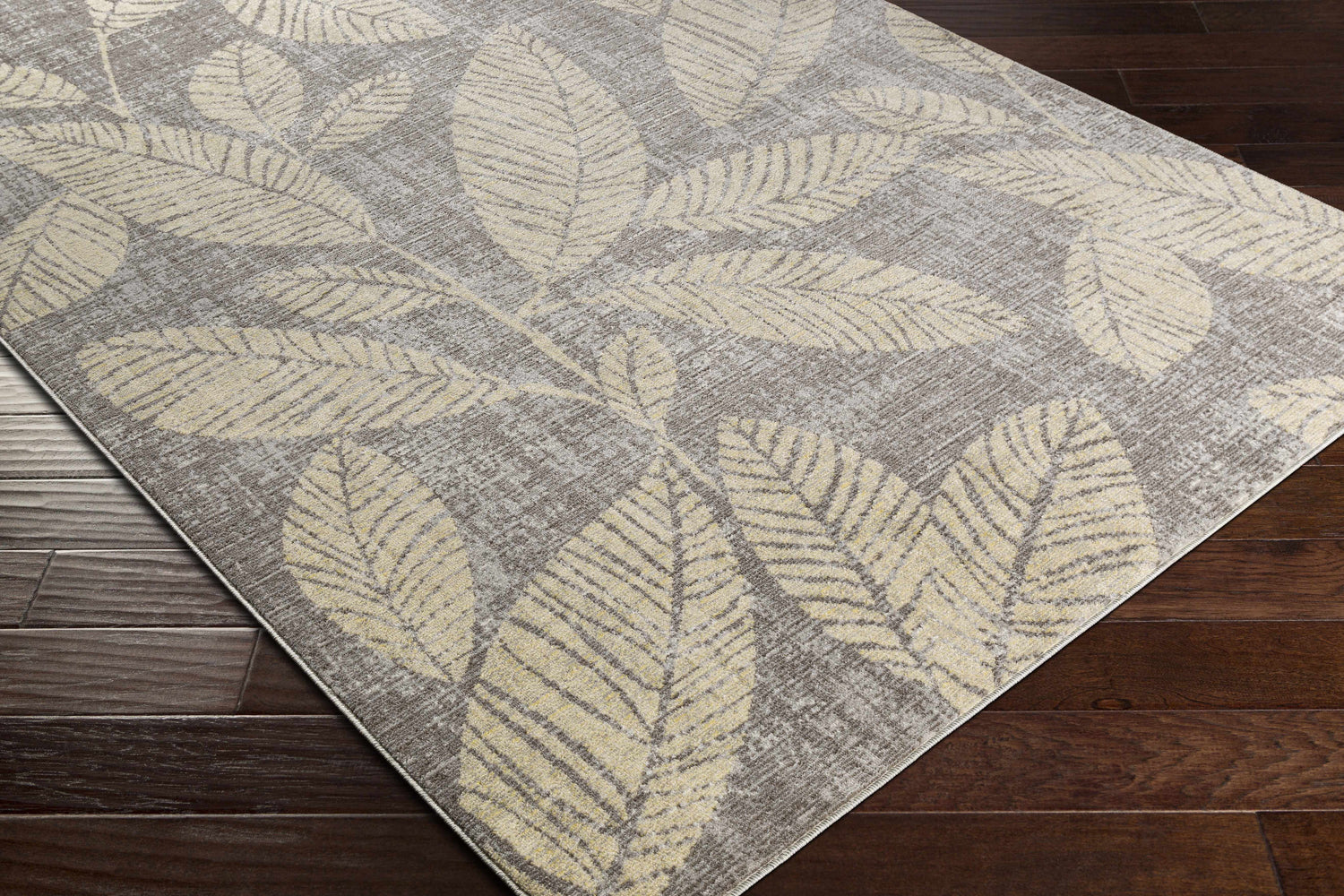 Close-up of the Snook Area Rug showing beige leaf pattern on a gray background, displayed on dark wood flooring. Corner
