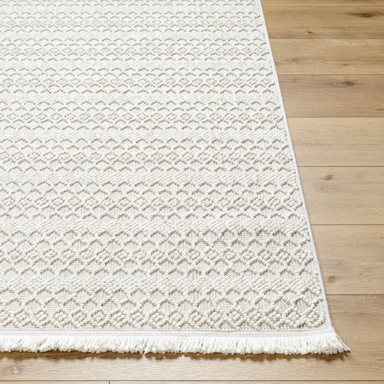 Close-up of the Hamal Area Rug in off white with a geometric pattern and fringe, displayed on a light wood floor. Front