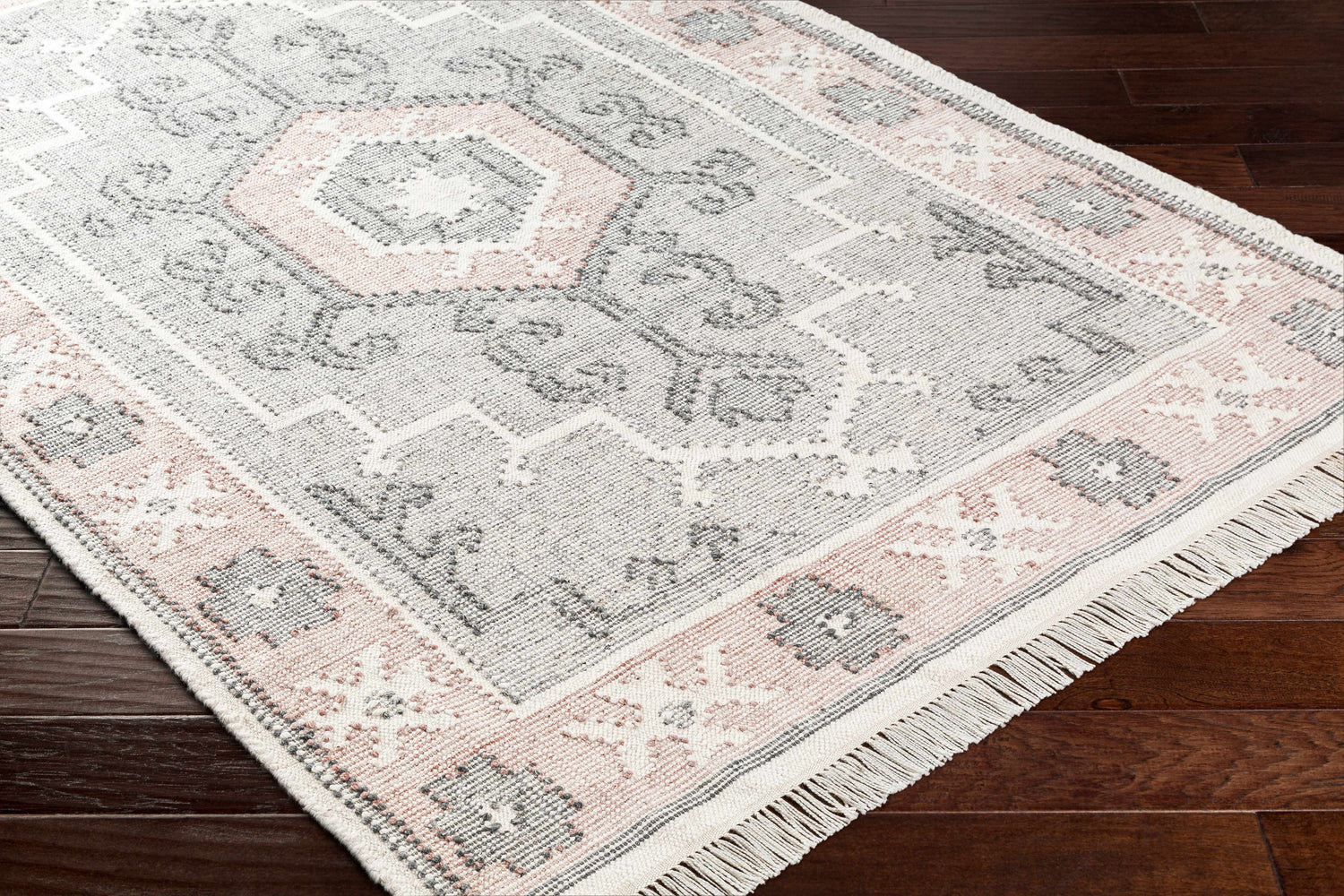 Close-up of the hand-woven Sosa Area Rug. Charcoal, cream, dusty pink, and light gray traditional design with fringe. Corner
