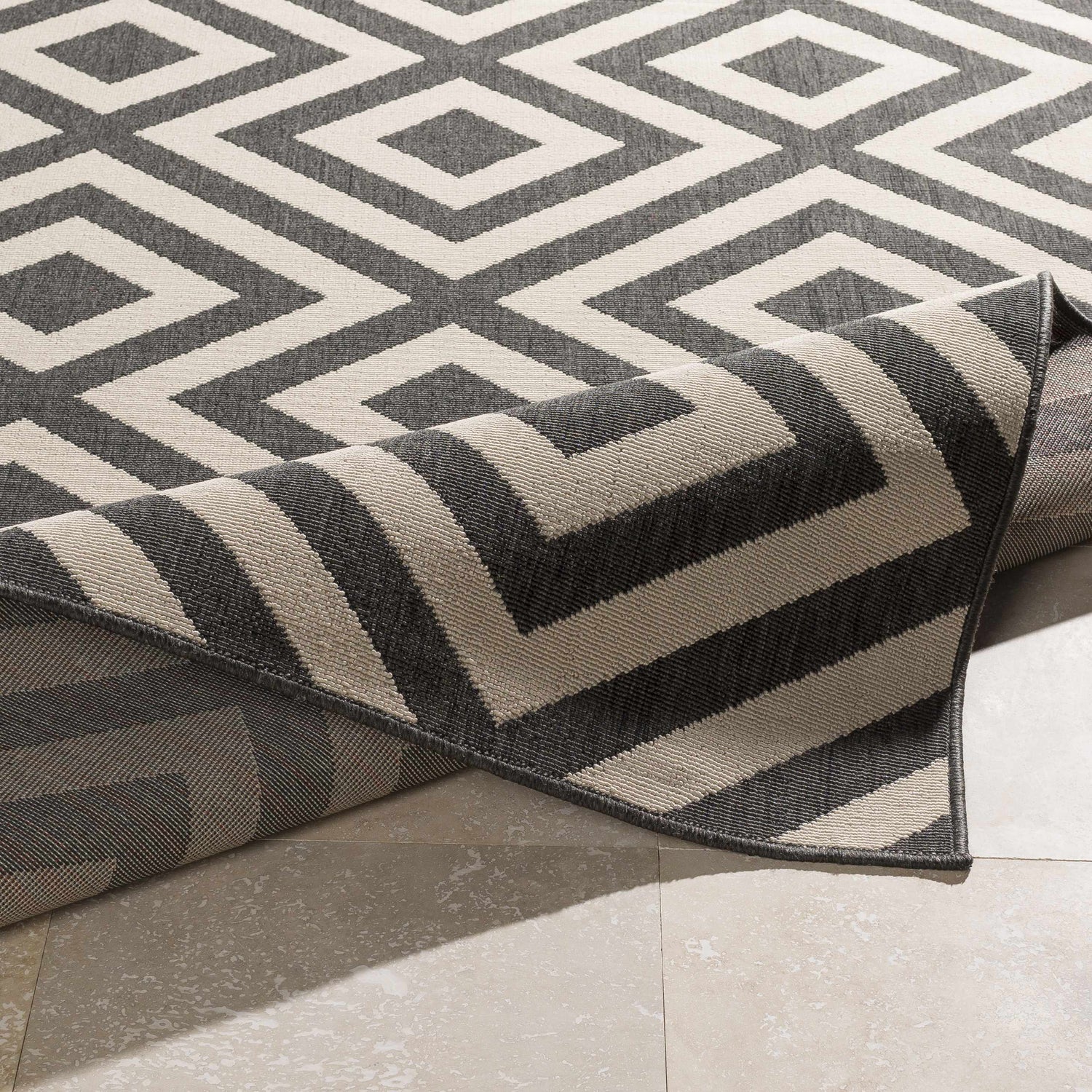 Close-up of Spilsby Geometric Outdoor Rug. Black and light beige diamond pattern, machine woven texture. Fold
