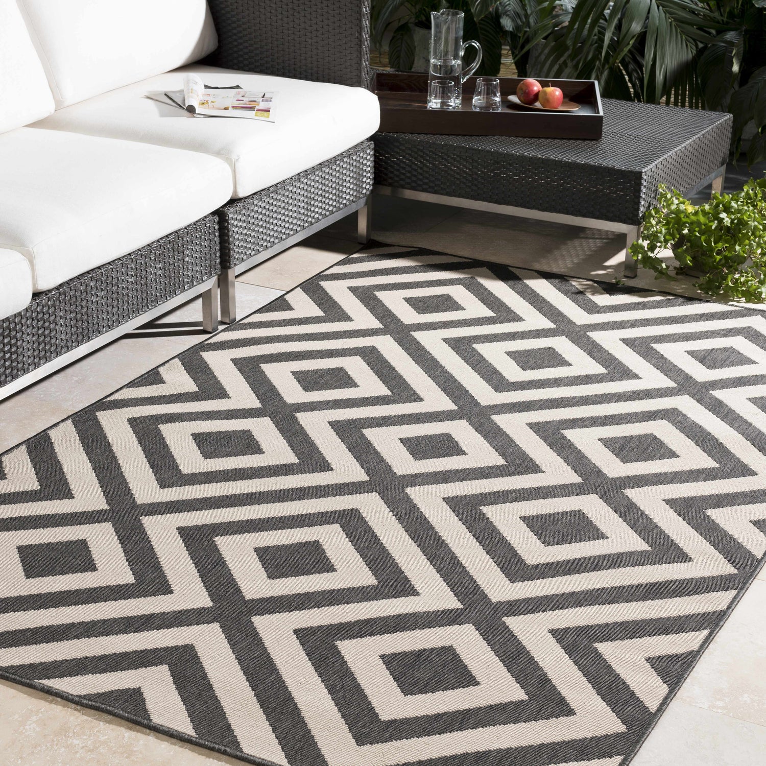 Outdoor patio scene with a black and light beige Spilsby Geometric Outdoor Rug, white couch, and woven coffee table. Roomscene-Rectangle
