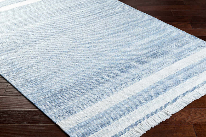 Close-up of the hand-woven Loic Silver Blue Area Rug. Features a striped pattern and fringed edge. Gray, blue, and white hues. Corner