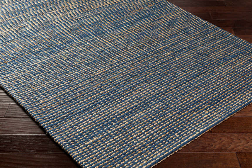 Close up of the hand woven Riki Navy Area Rug showing beige and navy blue jute texture on dark wood floor. Corner