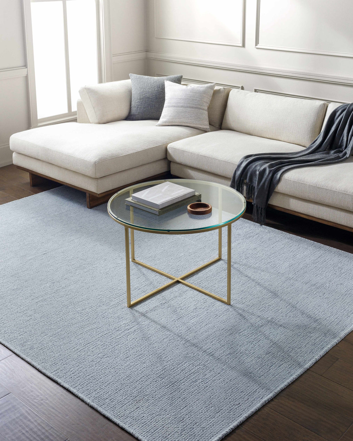 Living room scene with a light blue Isako Washable Rug, white sectional sofa, and gold-legged glass coffee table. Roomscene-Rectangle