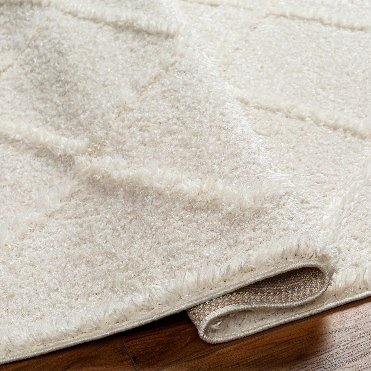 Close-up of the plush, cloud gray Daube Area Rug with a subtle geometric pattern and rolled edge detail on a wood floor. Fold