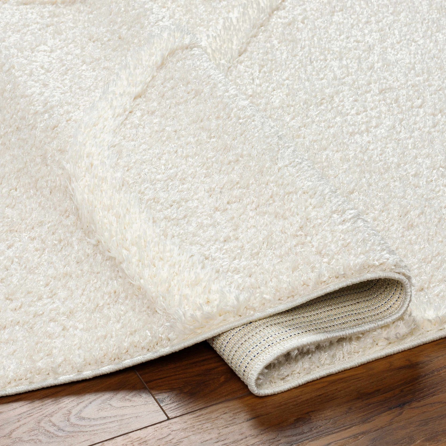 Close-up of the Horst Off White Area Rug showing its plush, shag texture and durable backing on a wood floor. Fold