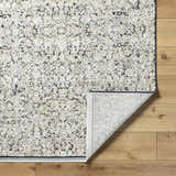 Close-up of the Haran Cloud Gray Area Rug showing its textured, abstract pattern and folded corner on a wood floor. Fold
