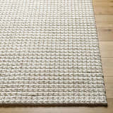 Close-up of the hand woven Lenci Area Rug in beige and rose cream, showing wool texture and braided design on wood floor. Front