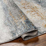 Close-up of the Adam Gray Blue Abstract Area Rug showing its gray, blue, and gold abstract design and soft, textured pile. Fold