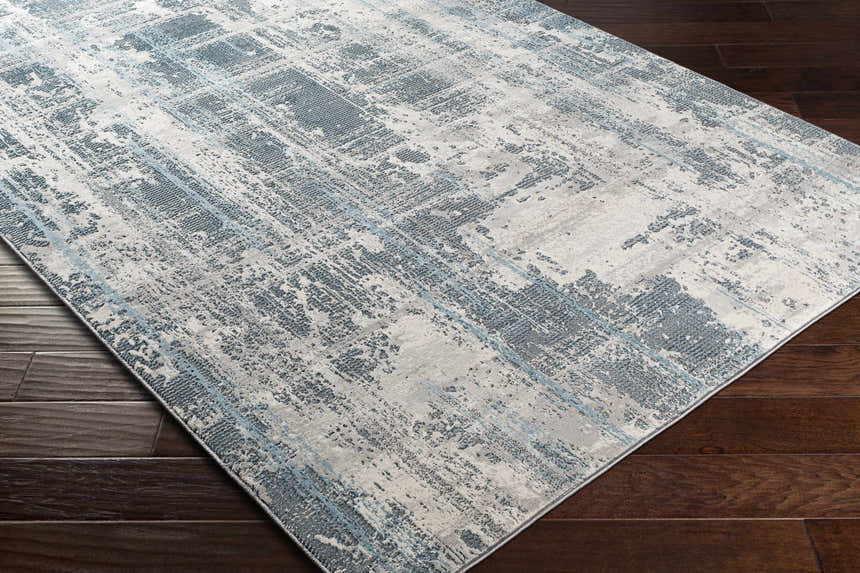 Close-up of the Stokenchurch Area Carpet showing a contemporary blue and gray abstract design on a dark wood floor. Corner