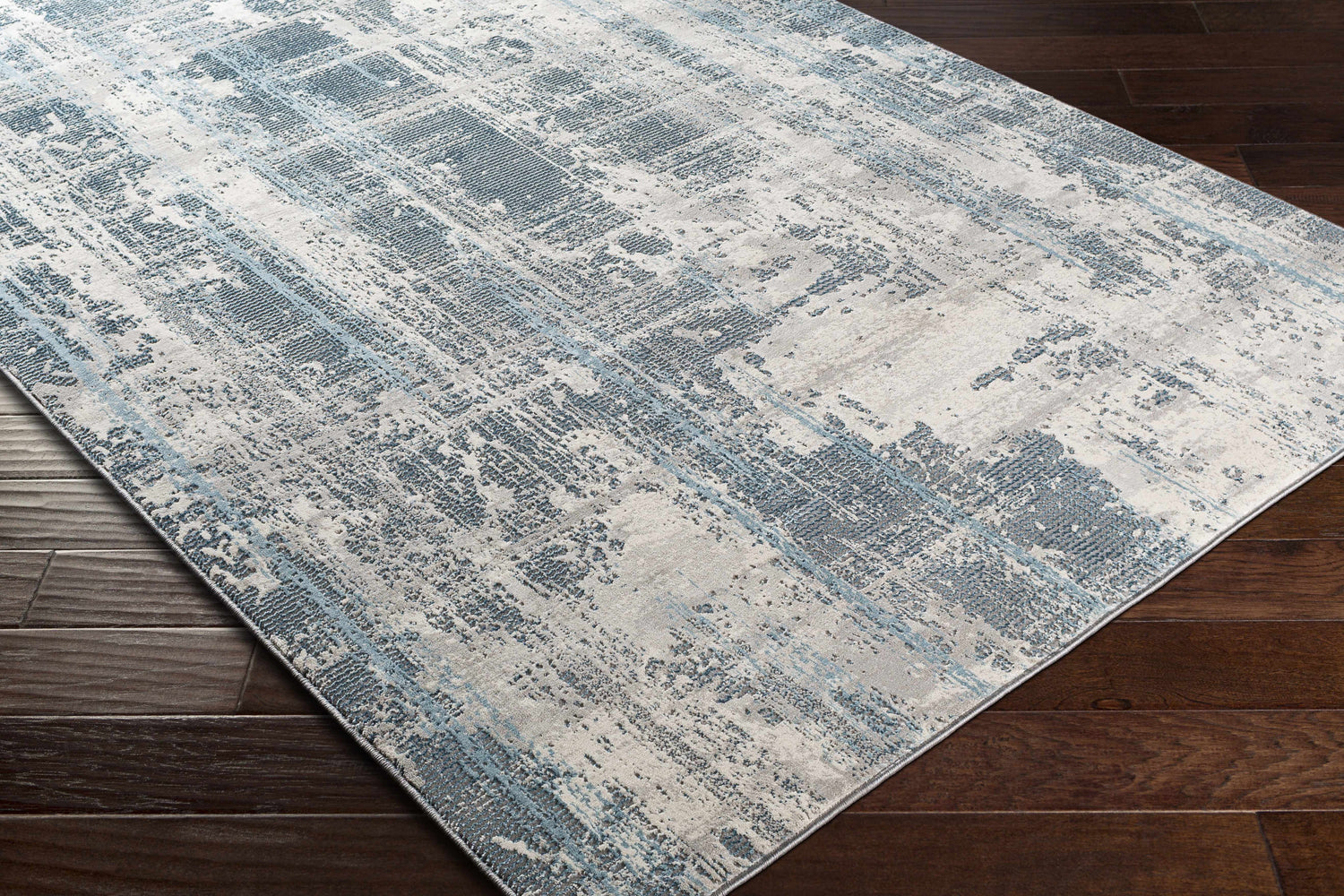 Close-up of the Stokenchurch Area Carpet showing a contemporary blue and gray abstract design on a dark wood floor. Corner