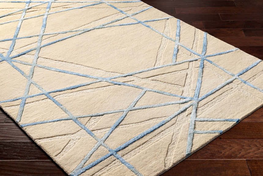 Close-up of Hiba Area Rug. Beige wool rug with a geometric slate blue line pattern. Hand-tufted texture. Corner