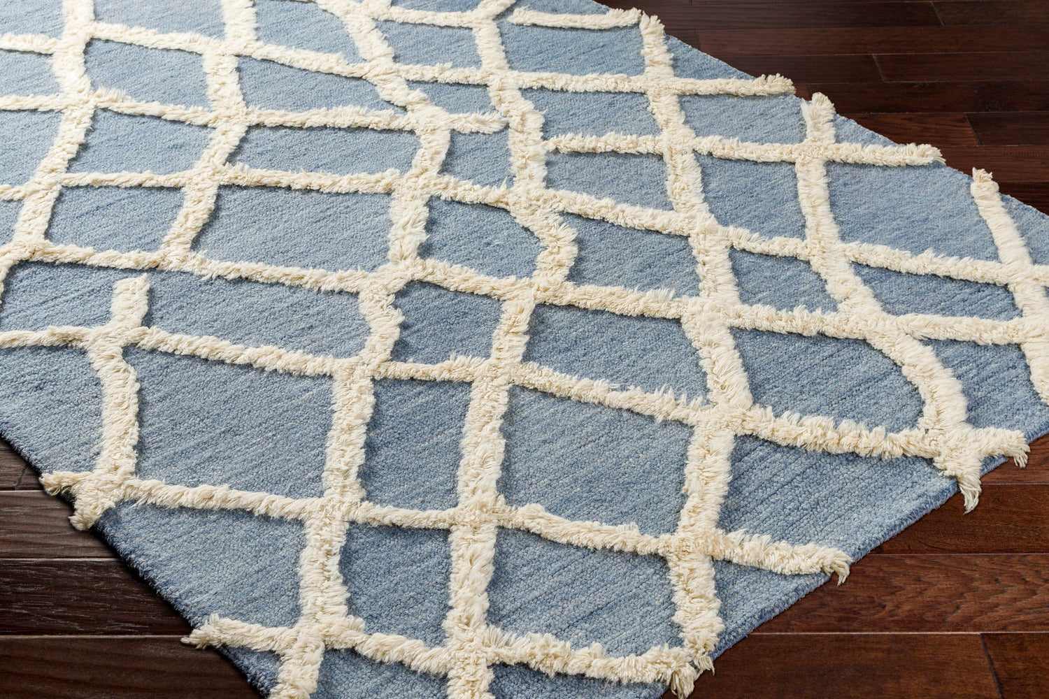 Close-up of Adica Blue Wool Area Rug showing geometric trellis pattern and soft, textured wool on dark wood floor. Corner