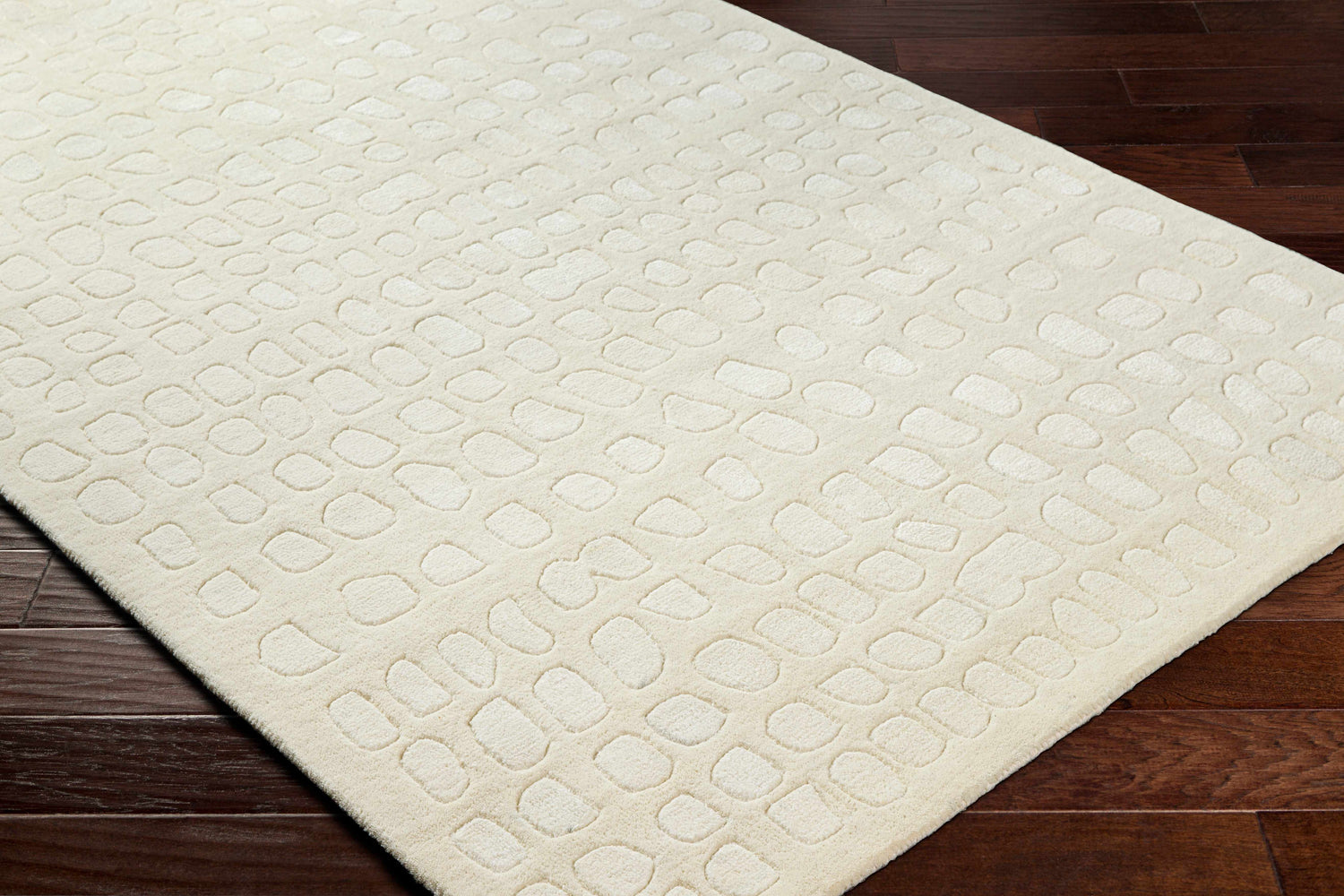 Close-up of the Arjan Cream Area Rug showing its textured, geometric pattern and soft, neutral color on dark wood flooring. Corner