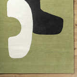 Close-up of the hand-tufted Terah Olive Area Rug. Features jet black, light silver, and moss abstract shapes on a prairie dust background. Corner
