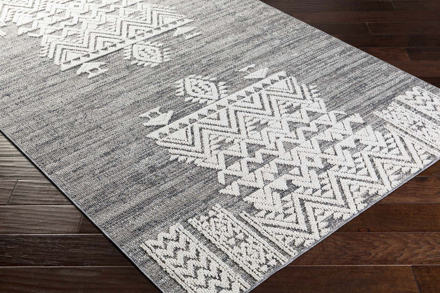Close-up of the Thorntonville Outdoor Rug. Gray rug with a white geometric, bohemian pattern on dark wood floors. Corner