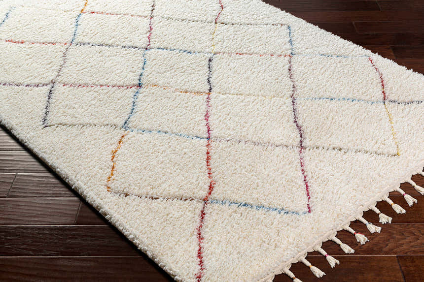 Close-up of the Tigao Area Rug on dark wood floor. Cream shag rug with black, orange, pink, and blue diamond pattern. Corner