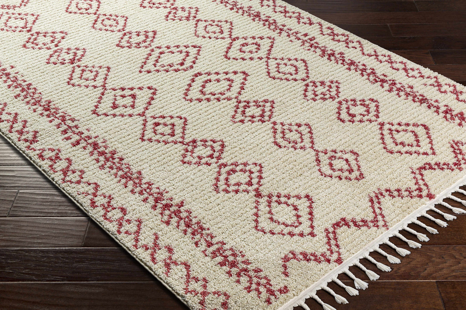 Close-up of the Tinawagan Area Rug. Cream rug with a red geometric pattern and tassels on dark wood flooring. Corner