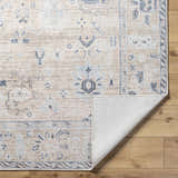 Close-up of the Toddy Area Rug, a beige rug with charcoal, mauve, and slate blue traditional patterns. Shows rug backing. Fold