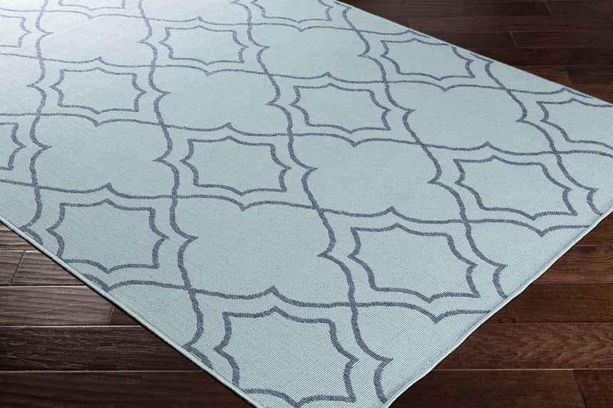 Close up of the Ursa Area Rug in pale blue with a navy quatrefoil pattern on dark wood flooring. Corner