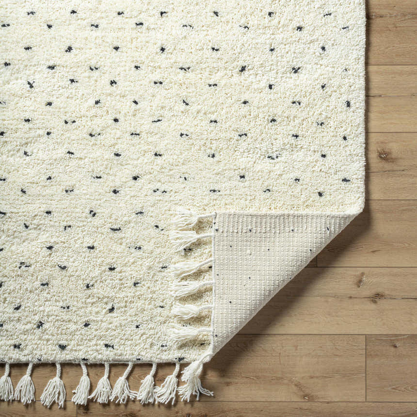 Close-up of the Yuuna Area Rug. Pearl wool rug with small black dots and tassels on a wood floor. Fold