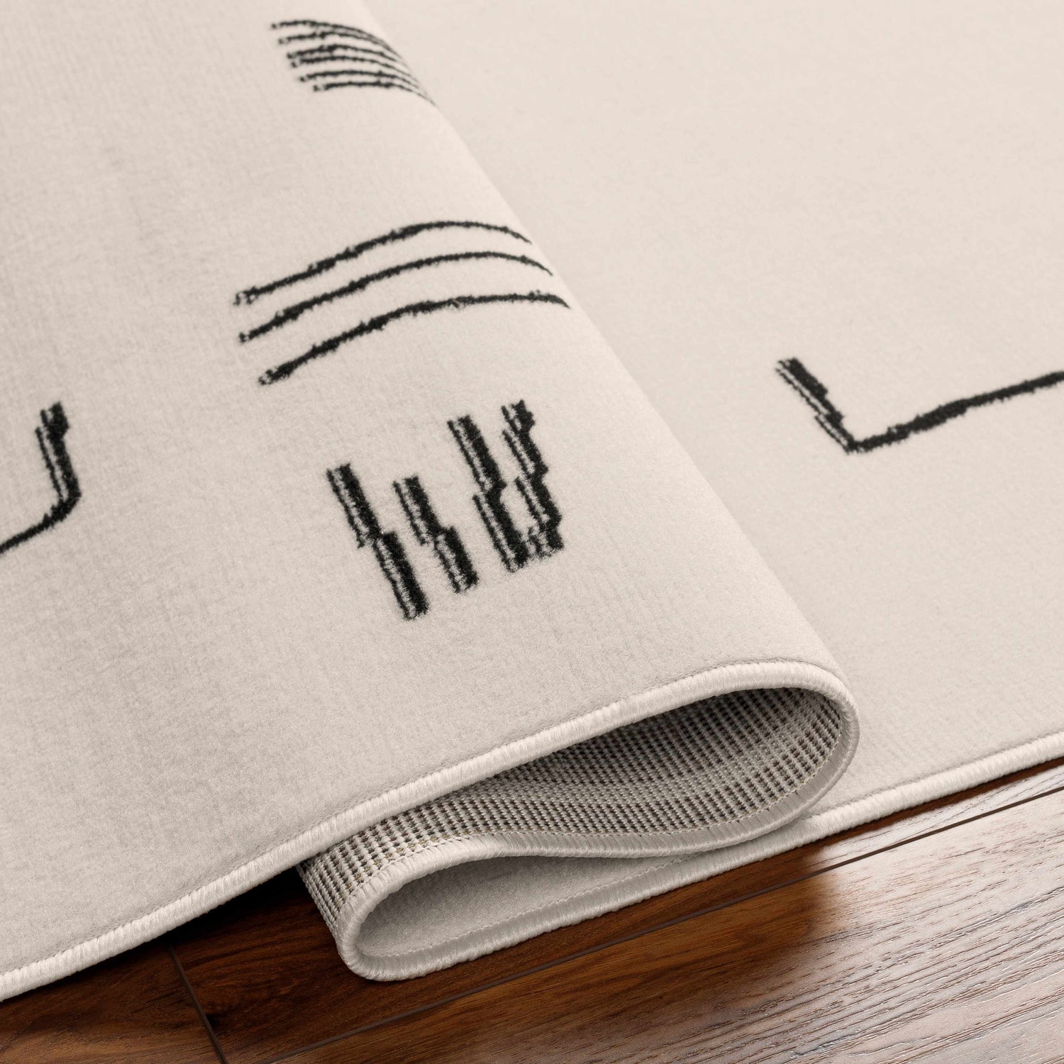Close-up of the Zale Area Rug showing its bone color and dark gray abstract line pattern. Machine woven texture visible. Fold